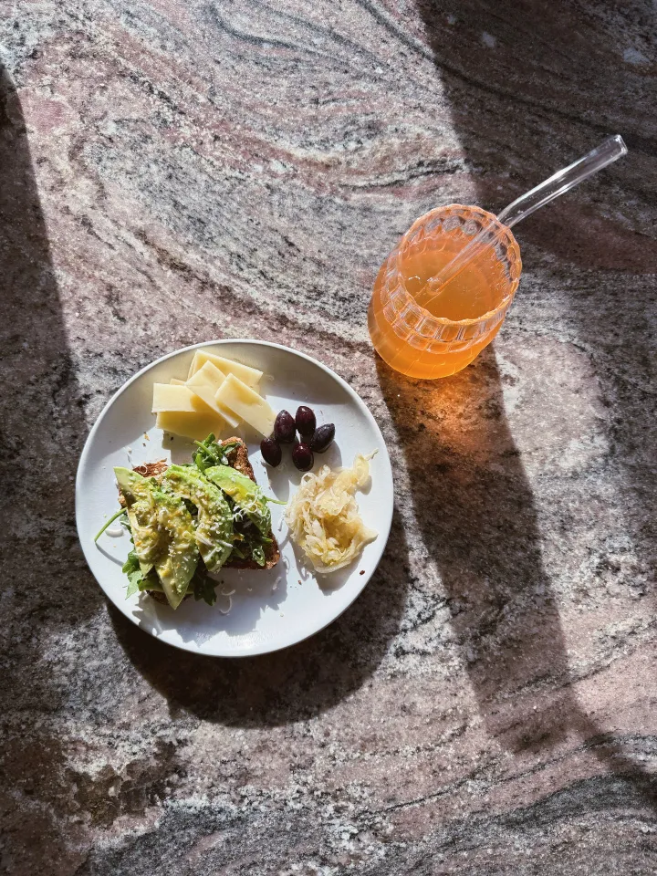 A top-down view of a small plate and cup. The plate is white ceramic and contains avocado toast, sliced low fat Swiss cheese, five dark olives, and a small amount of sauerkraut. The cup is clear and contains a low sugar peach clementine soda.