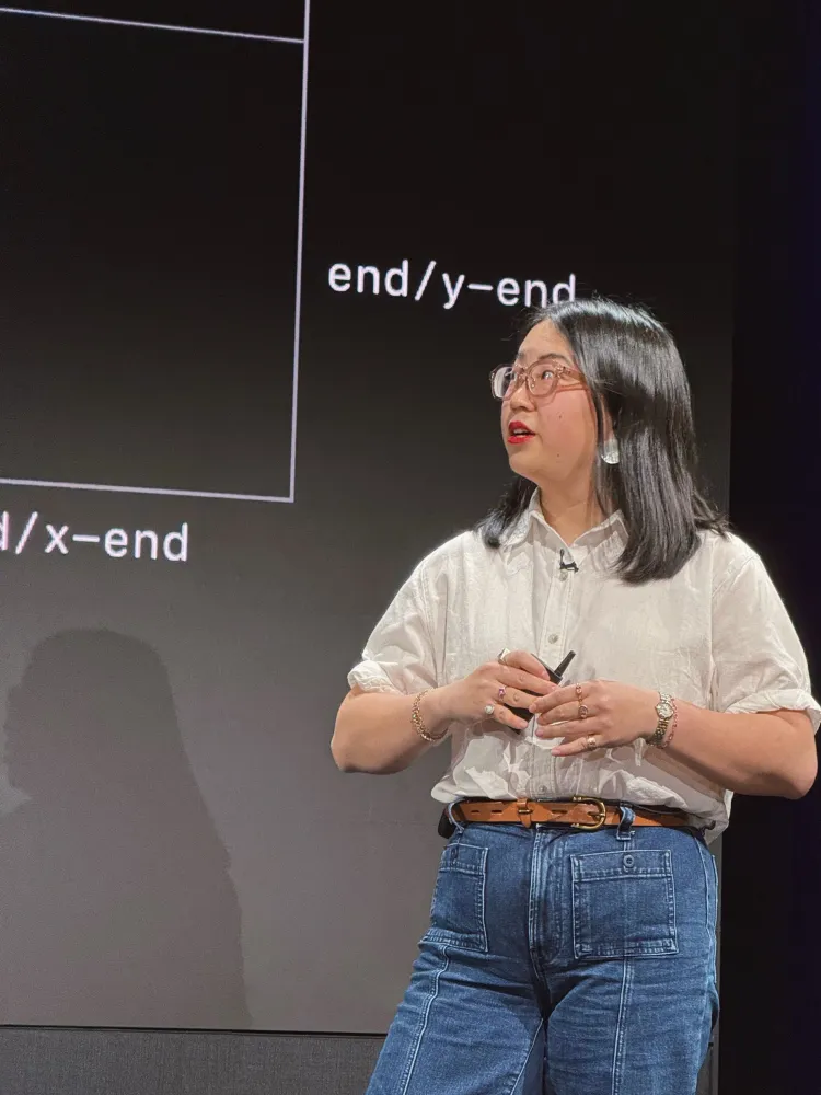An Asian woman giving a presentation about CSS features. She’s standing in front of a large projector screen with a graphic of CSS position-area and is wearing chunky acetate glasses, red lipstick, a white short-sleeved collared shirt, and dark blue jeans.