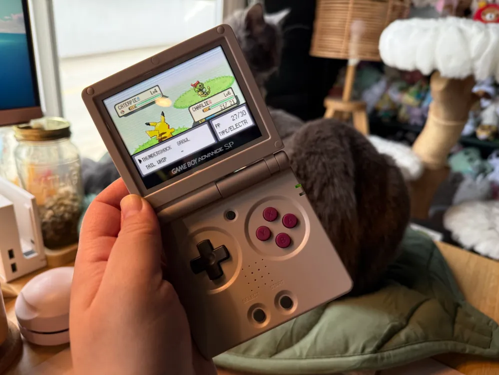 I’m holding up a GBA SP emulator device in front of the camera. The game being played is Pokémon Leaf Green. Behind the emulator are two grey cats sitting on a green leaf mat on top of a bamboo desk.