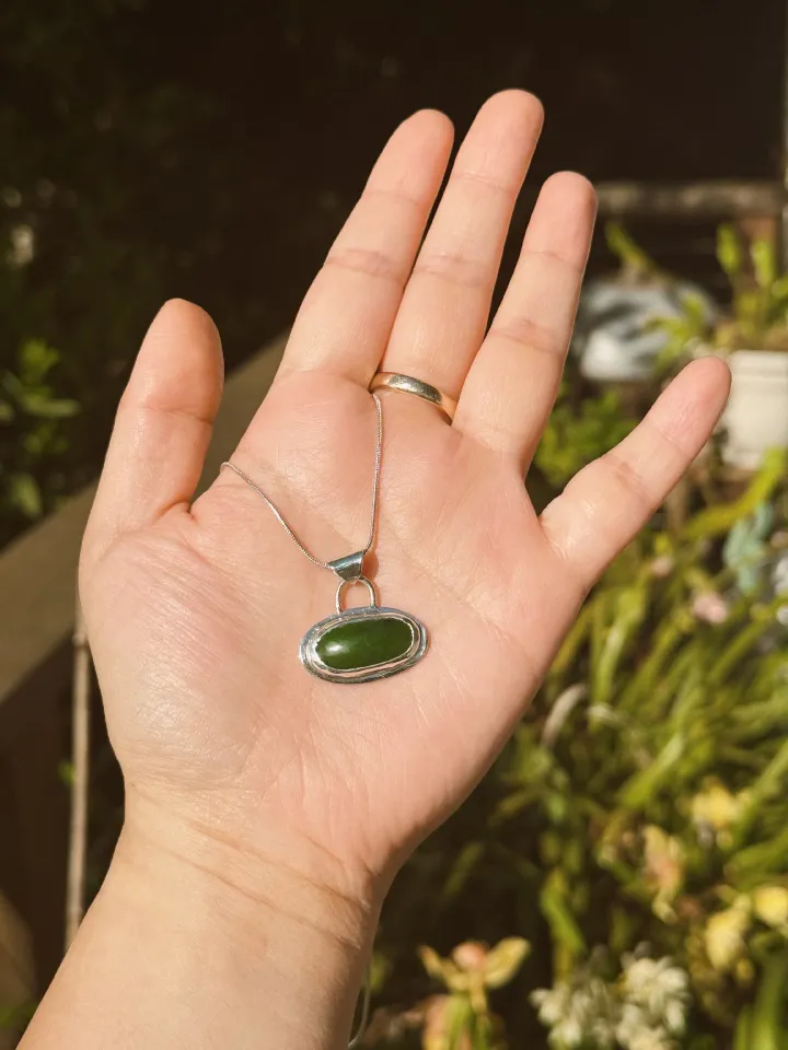 A elongated green jade cabochon set east-west in a sterling silver bezel pendant. The pendant is being held up on a silver chain in front of plants in the afternoon sun.