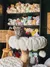 A grey kitten sitting on top of a fuzzy cat tree in front of a shelf filled with plushies and stuffed animals.