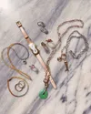 A collection of jewerly on a white marble background, including a slim dual-faced watch, pink sapphire rings, rainbow beads, and a bright green jade pendant