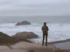 A man with his back to the camera, facing a rocky overlook and white ocean waves.