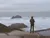 A man with his back to the camera, facing a rocky overlook and white ocean waves.