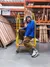 A person with long bleached hair and a bright blue jacket sitting on a yellow rolling ladder in front of wood supplies at Home Depot.