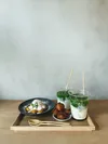 A carefully arranged spread of matcha lattes, kinako-dusted sticky rice balls, and baby castella on a wooden tray atop a wooden table with a minimalist concrete backdrop.