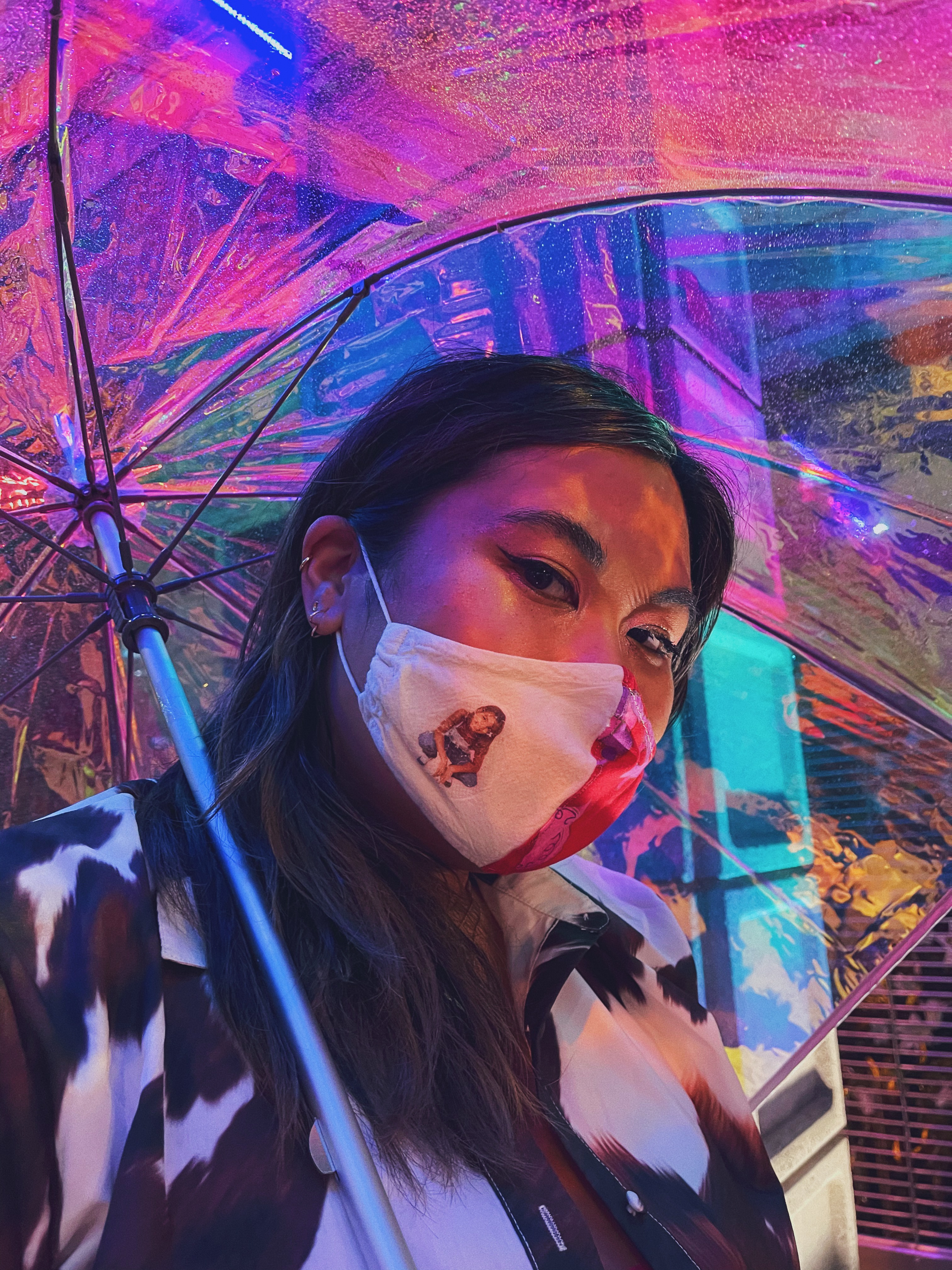 A person with long hair and a Britney Spears face mask holding an iridescent umbrells over their head.
