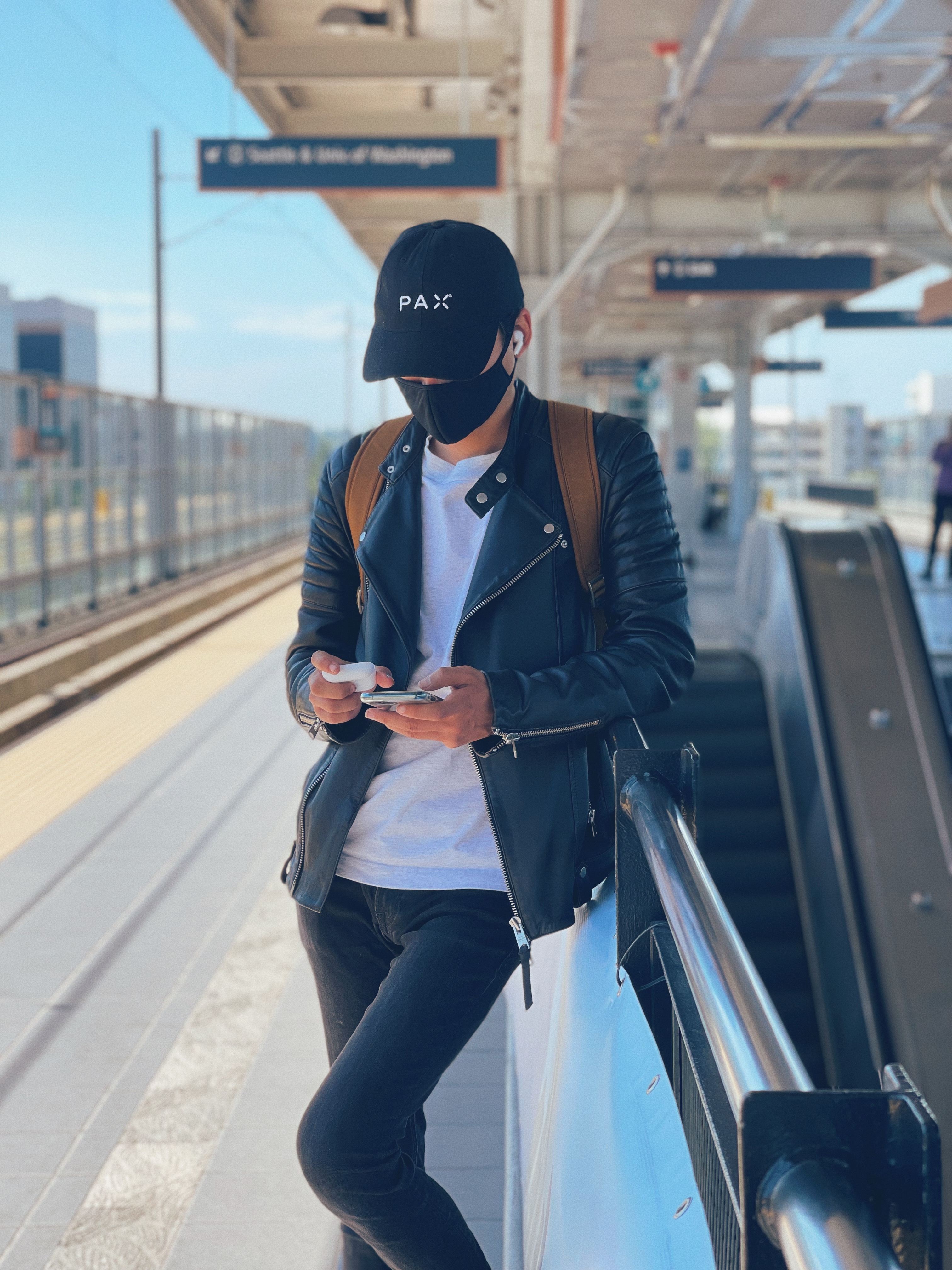 A man in a leather jacket, black hat, and black mask looking at his phone and earbuds leaning against a train station wall in SeaTac, Washington.