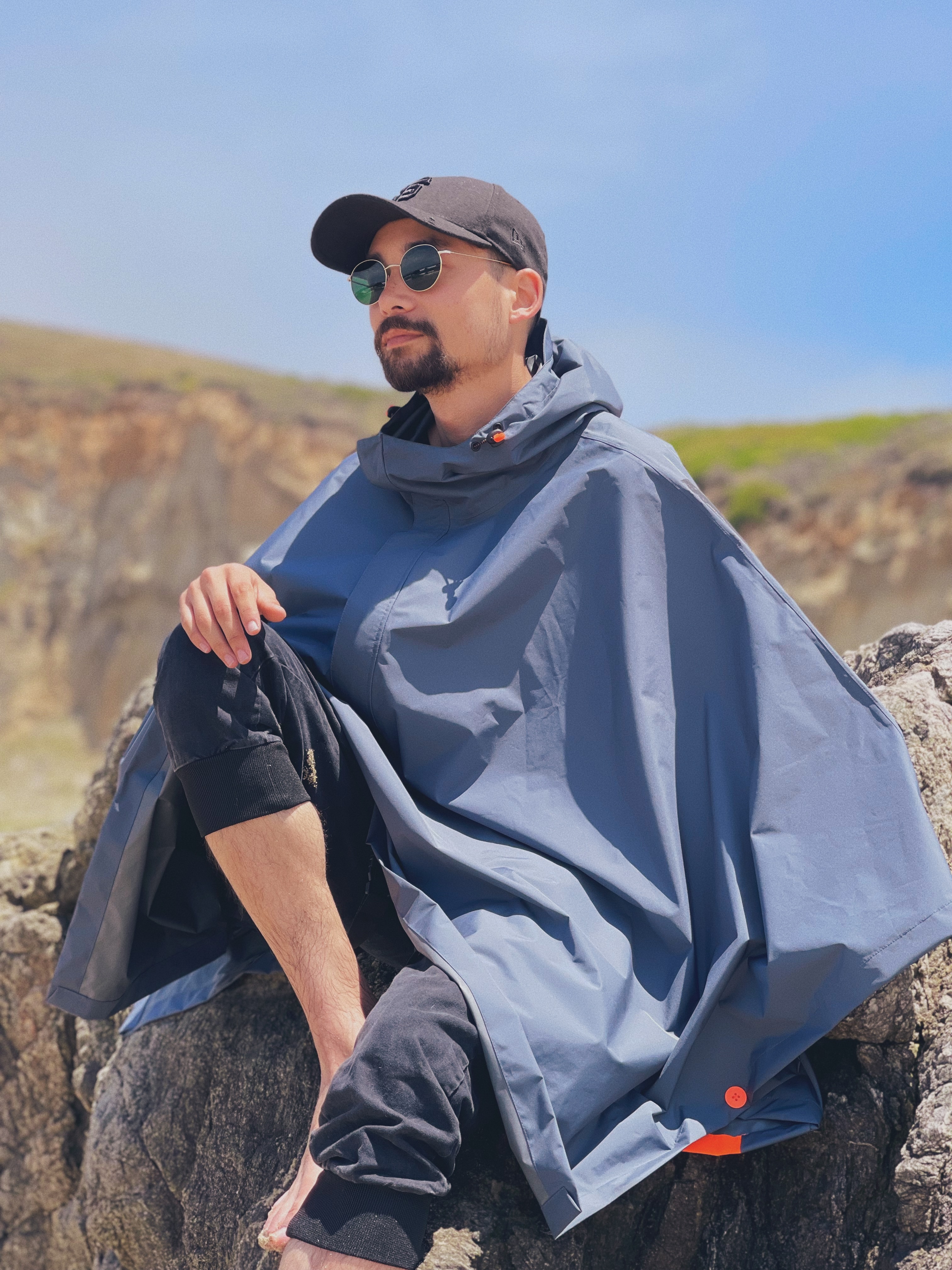 A man in a waterproof poncho, sunglasses, and a hat sitting on a rock on Kehoe Beach.
